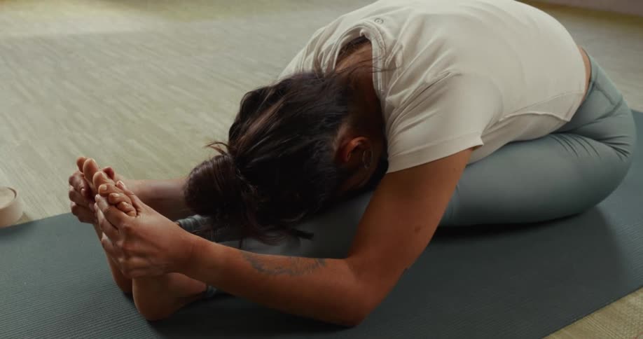 A flexible woman in a white t-shirt holds a deep seated forward bend on her yoga mat