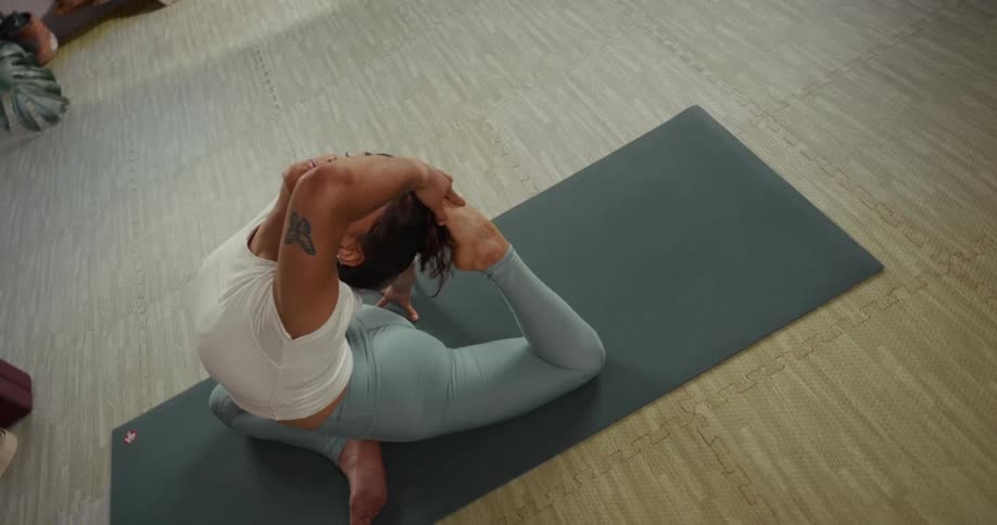 A high-angle view follows a flexible woman as she holds an advanced King Pigeon yoga pose on a mat in a serene studio