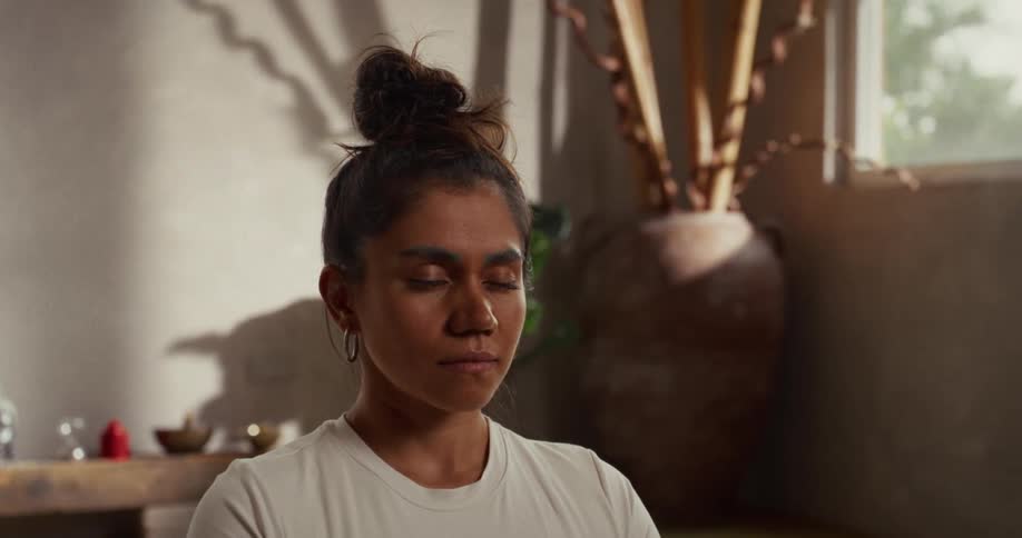 A serene woman with her hair in a bun meditates peacefully as incense smoke gently wafts past her face in a sunlit room