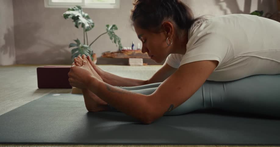A flexible woman performs a seated forward bend yoga stretch on a mat in a minimalist studio