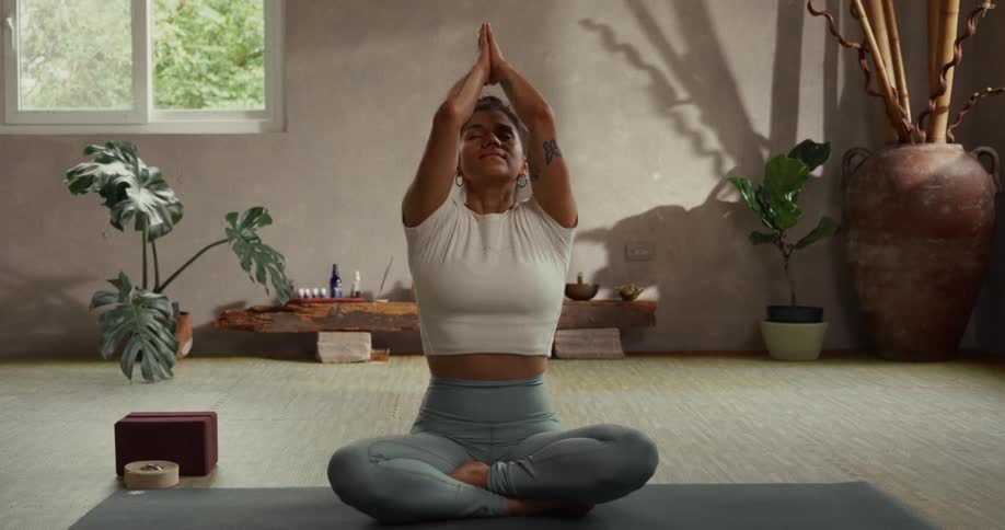 A calm woman with closed eyes meditates in a lotus position, slowly raising her arms and bringing her hands together in a prayer pose in a peaceful studio