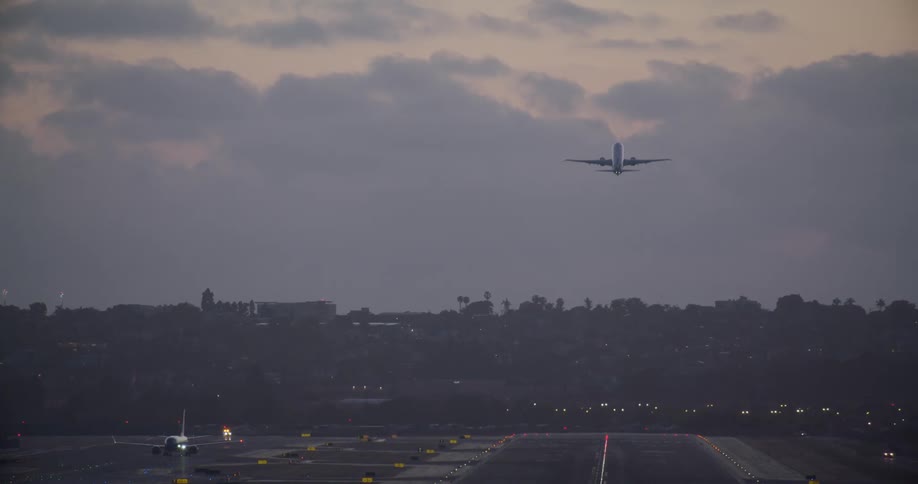 A passenger jet takes off from a busy airport runway at dusk as another plane taxis in the foreground