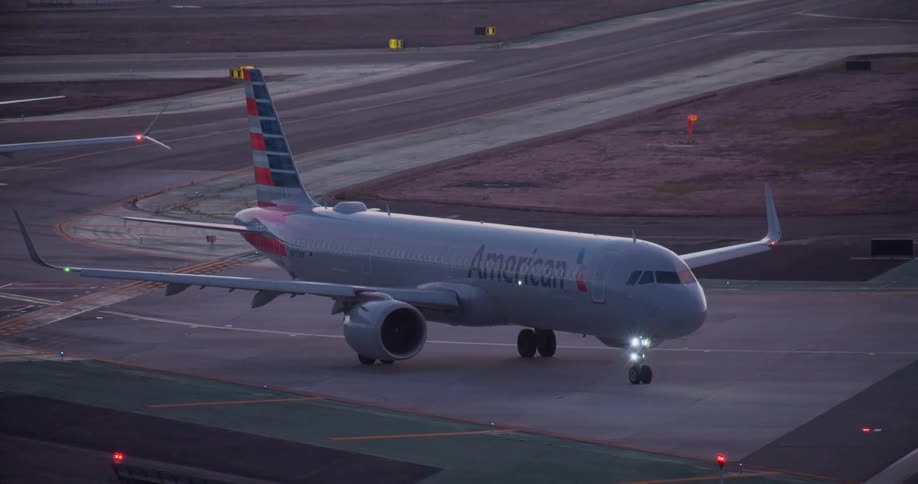 An American Airlines airplane with flashing lights waits on the tarmac at dusk as an Alaska Airlines jet taxis in the background