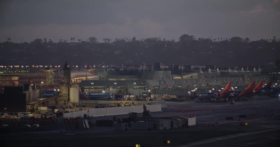 An airport terminal with planes parked at the gates is brightly illuminated against the dark evening sky