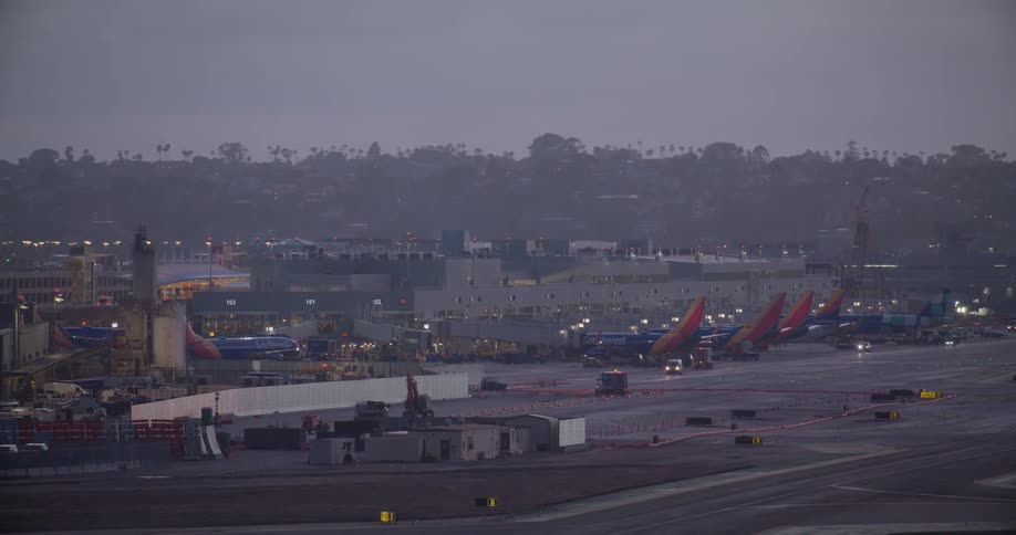 A wide shot of a busy airport tarmac at dusk with airplanes parked at the terminal and ground vehicles moving about
