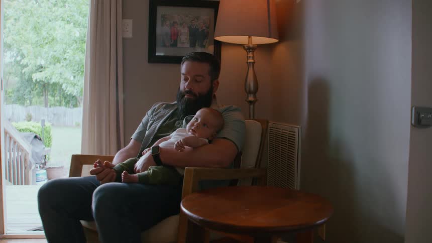 A bearded father peacefully sits in a chair in his home, holding his newborn baby in his arms