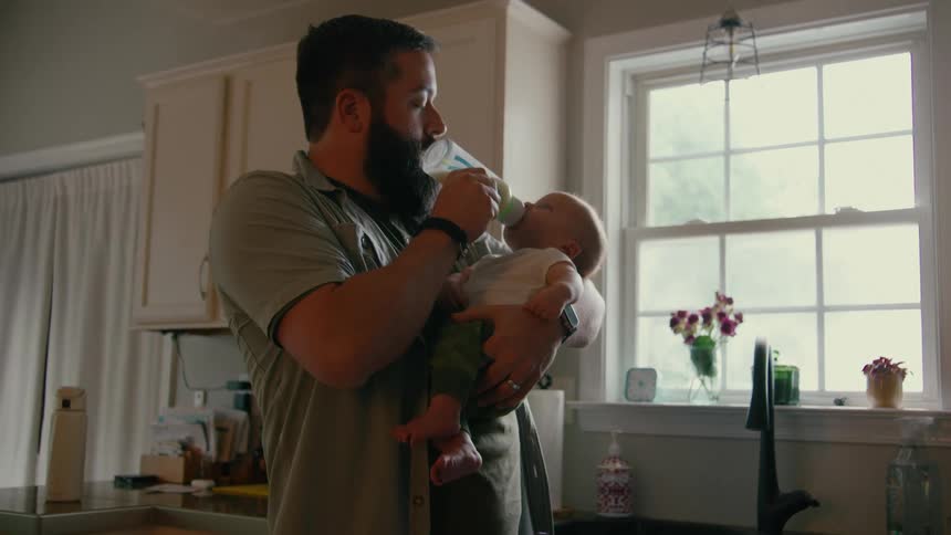 A bearded father lovingly feeds his newborn baby with a bottle while holding him in his arms in the kitchen