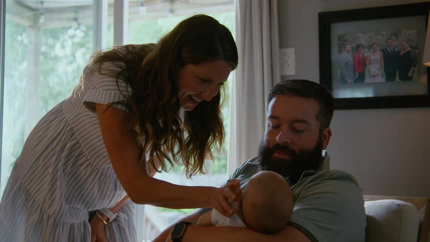 A happy mother smiles while gently cleaning her newborn baby, held in the father's arms