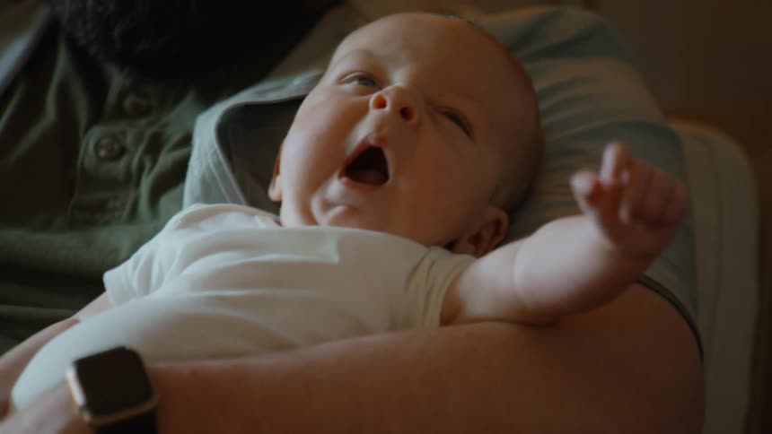 A close-up of a cute newborn baby yawning and stretching in slow motion while being held in their father's arms