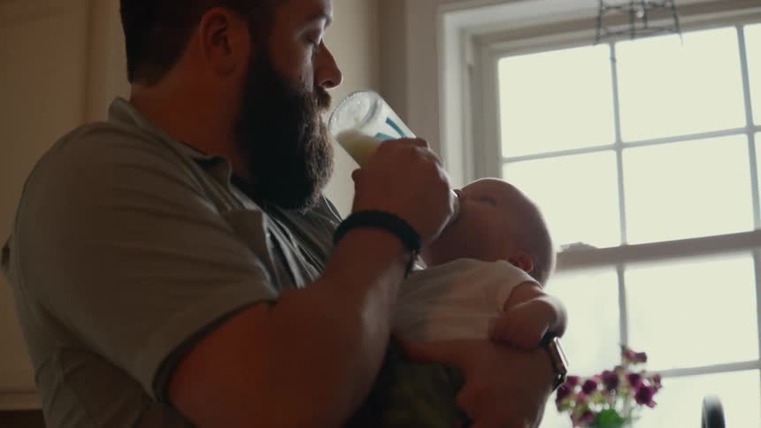 A bearded father lovingly holds and bottle-feeds his newborn baby in a naturally lit kitchen