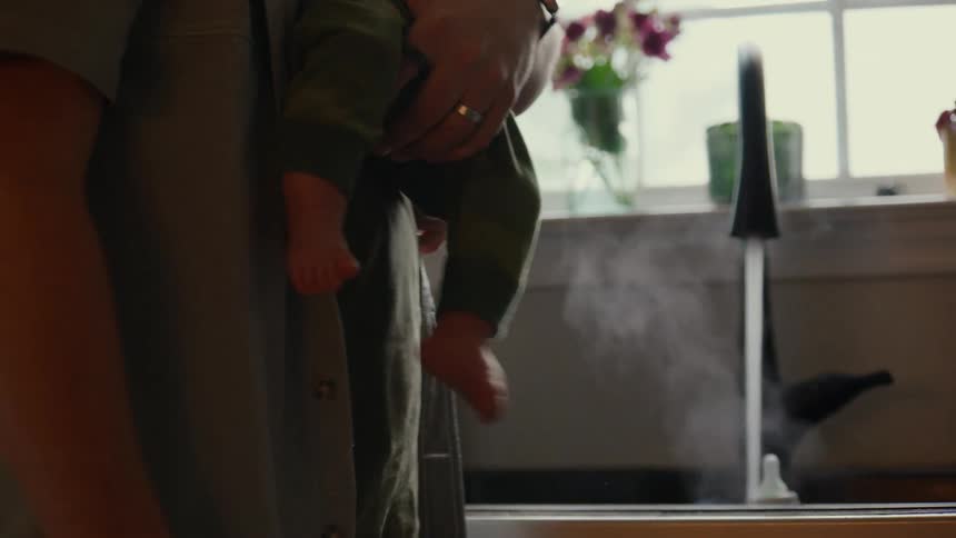 A father multitasks by holding his baby while cleaning and sanitizing bottles in a steamy kitchen sink in slow motion