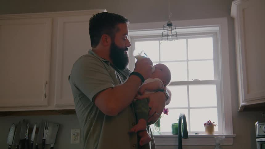 A new father with a full beard holds and lovingly feeds his newborn baby with a bottle in their bright kitchen