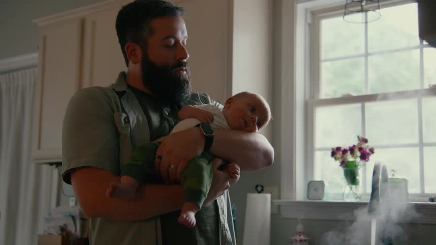 A new father with a full beard cradles his newborn baby in his arms while standing in the kitchen