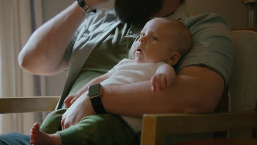 A father holds his curious infant baby in his arms while talking on the phone in their sunlit home