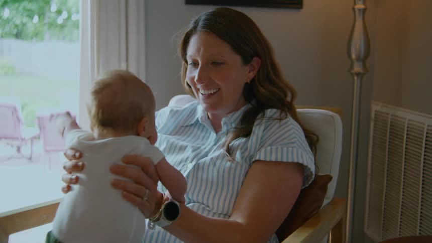 A joyful mother smiles and laughs while holding her newborn baby on her lap in a sunlit room