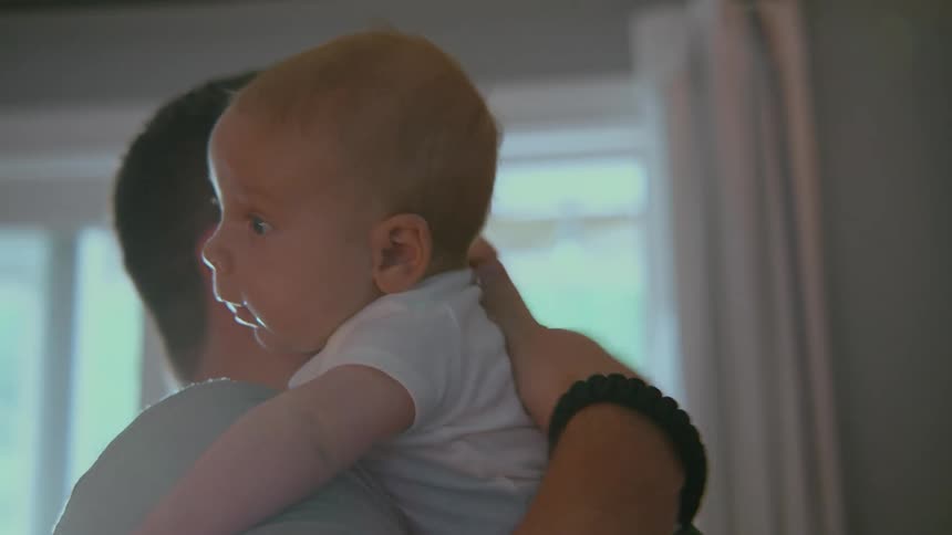 A father gently comforts his newborn baby, holding them over his shoulder and patting their back in a softly lit room