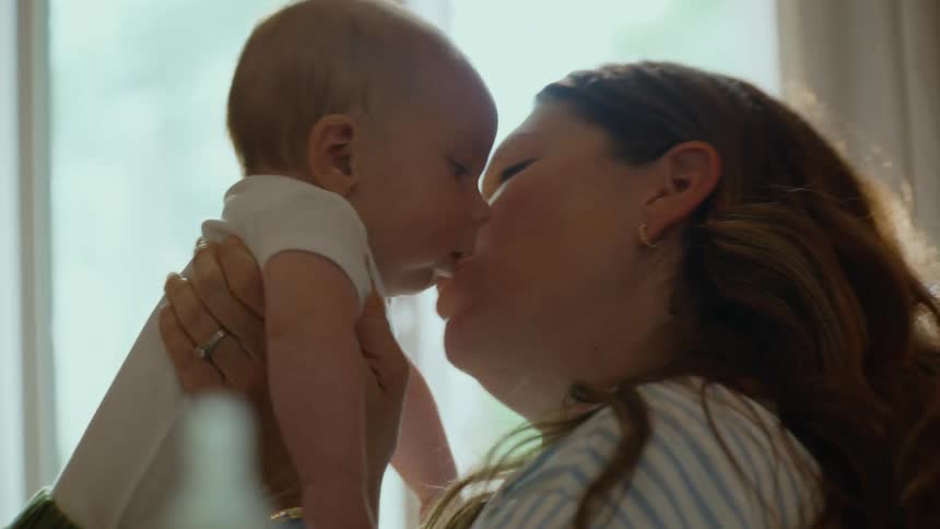 A loving mother holds up her newborn baby, showering it with kisses and affection in a sunlit room