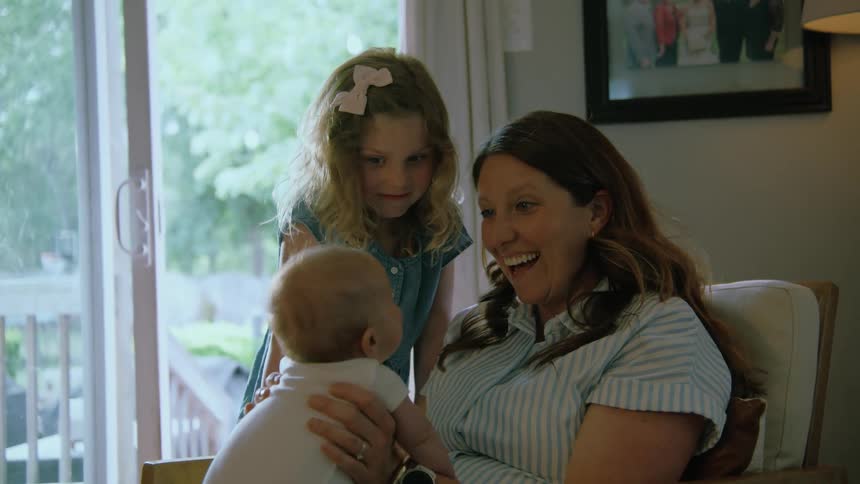 A smiling woman holds a baby while a young girl looks on lovingly in their cozy living room