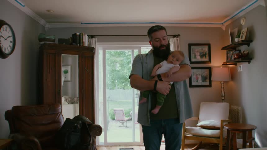 A bearded father lovingly holds his newborn baby in his arms while standing in their cozy living room