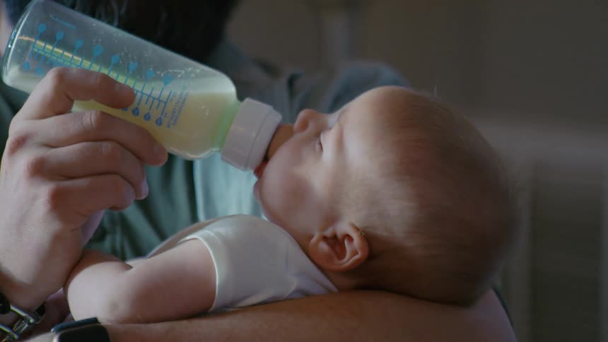 A close-up view of a father tenderly bottle-feeding his sleepy newborn baby