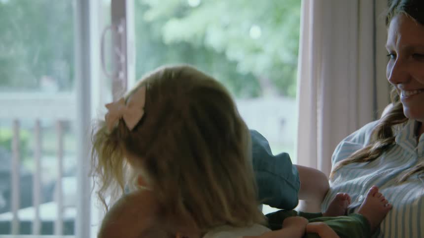A sweet older sister gently kisses and hugs her newborn sibling held by their loving mother