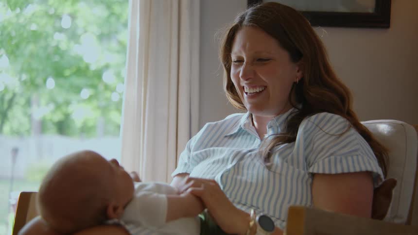 A joyful new mother smiles and laughs as she holds her newborn baby on her lap in a sunlit room
