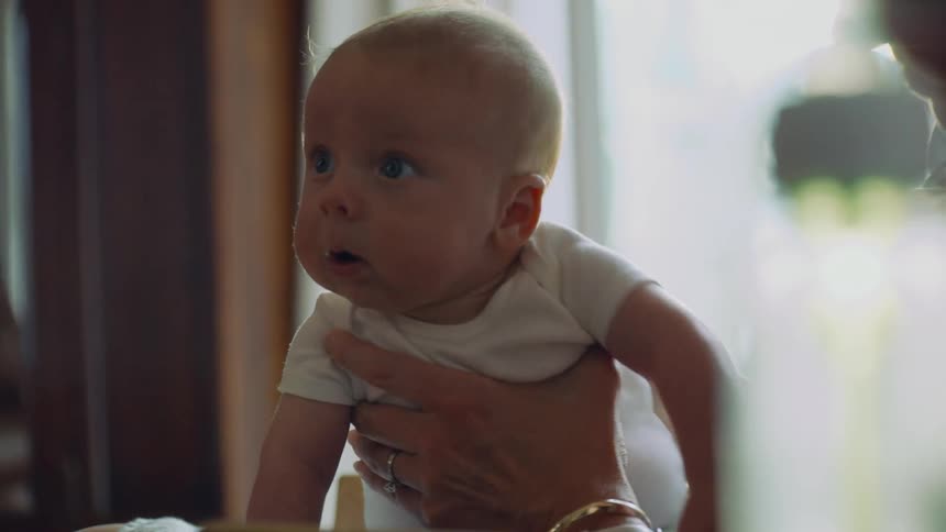 An adorable newborn baby with chubby cheeks and wide eyes is held up during tummy time in a warmly lit home