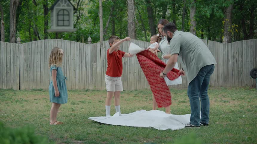 A happy family with three children lays out a blanket for a picnic in their backyard in slow motion