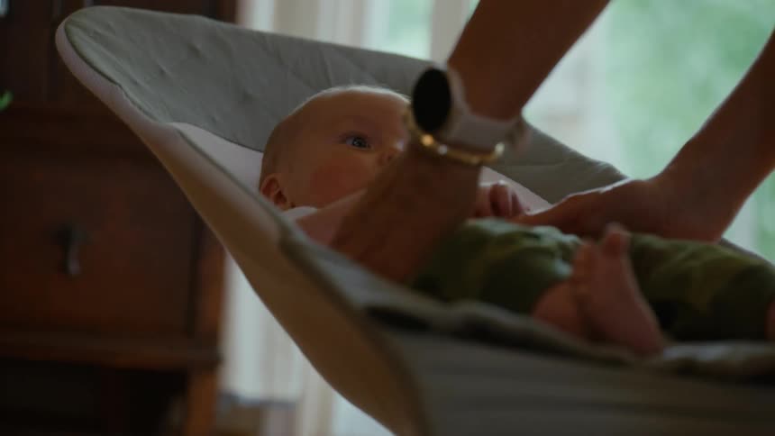 A mother gently picks up her newborn baby who is resting in a bouncer chair inside their home