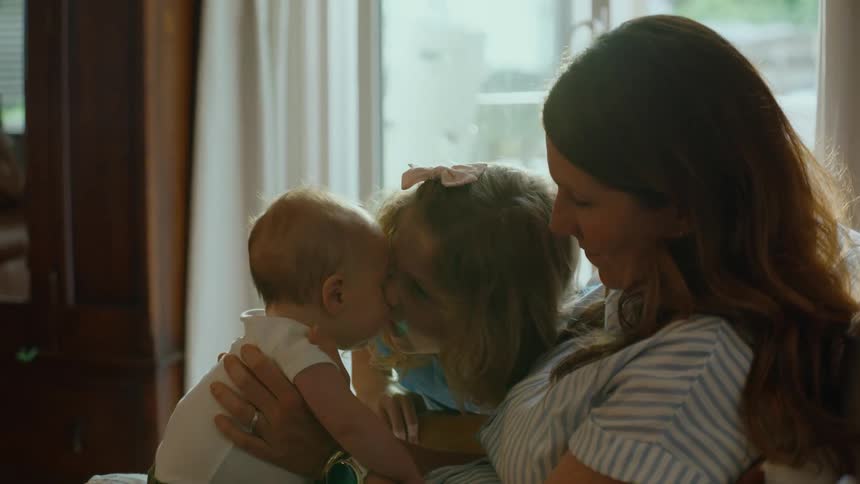 A young girl gently kisses her baby sibling on the forehead as their loving mother holds the baby in her arms