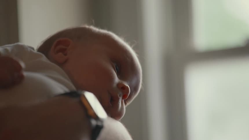 A sleepy newborn baby rests peacefully on their parent's shoulder in a softly lit room