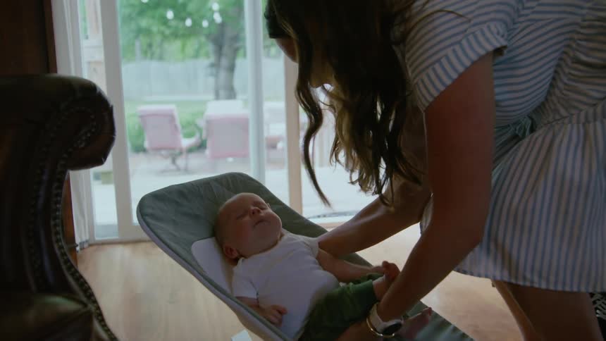 A loving mother kisses her newborn baby and gently places him to sleep in a bouncer as the father looks on