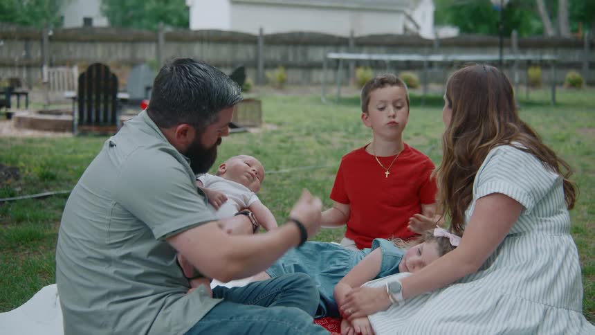 A family of five shares a tender moment while picnicking on a blanket in their grassy backyard