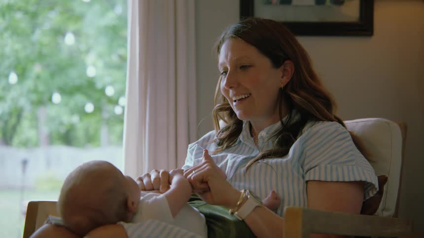A new mother lovingly plays with her newborn baby's hands and feet while holding them in a chair by a window