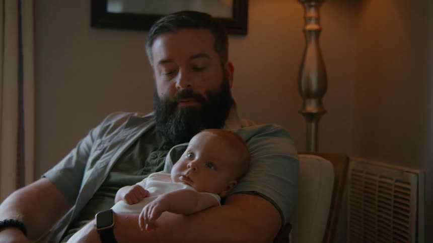 A bearded father sits in a softly lit room, holding his newborn baby in his arms