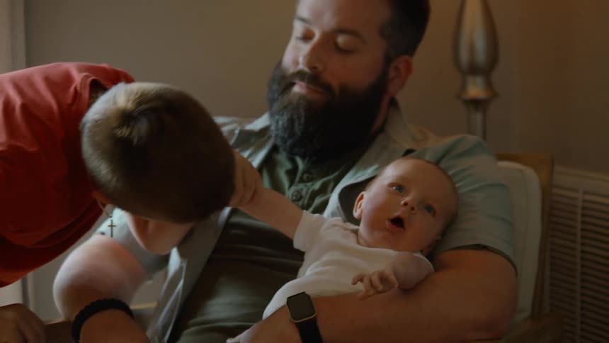 A gentle older brother kisses his newborn sibling's hand while their father holds the baby in a cozy living room