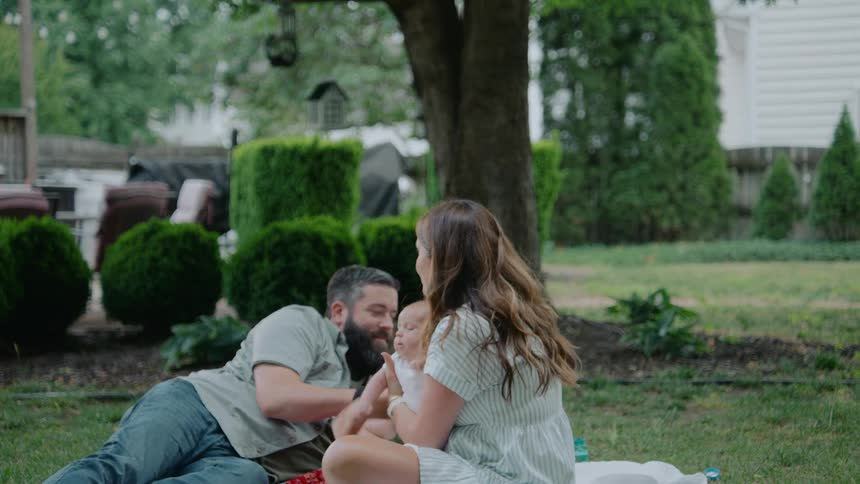 Parents sit on a blanket with their newborn baby as their energetic older children run around them in slow motion in the backyard