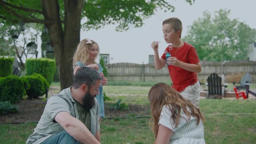 A loving family gathers in their backyard, with older siblings joyfully blowing bubbles around their parents and newborn baby
