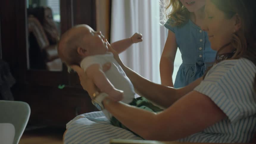 A young girl gently kisses her baby brother held in their mother's lap in a tender family moment