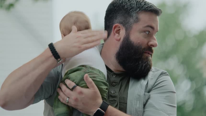 A bearded father lovingly holds and soothes his baby, patting their back while another child plays in the background