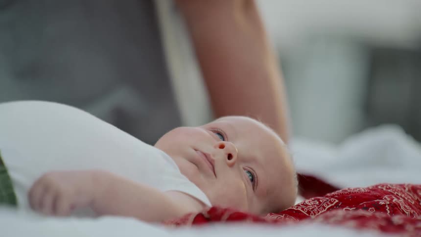 A curious newborn baby with blue eyes calmly lies awake on a bed as its mother watches in the background