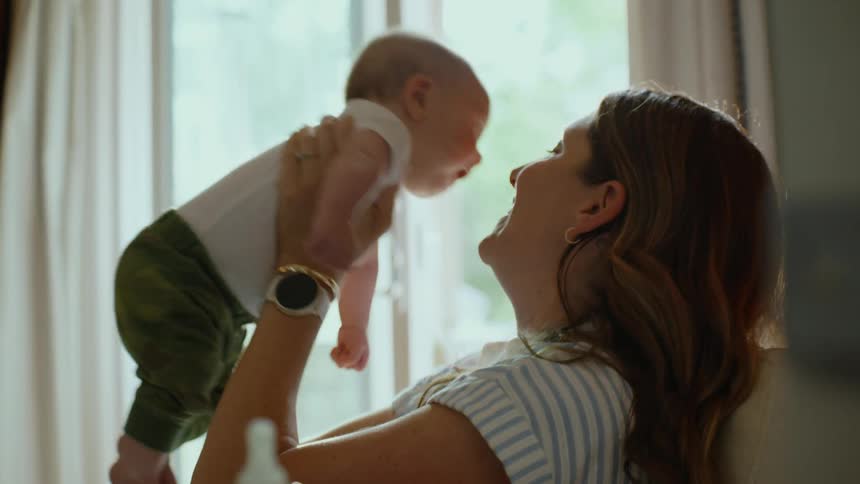 A loving mother joyfully lifts her baby in the air and gives them a kiss in slow motion