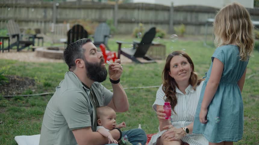 A happy young family with a baby and toddler daughter blow bubbles together while having a picnic in their suburban backyard in slow motion