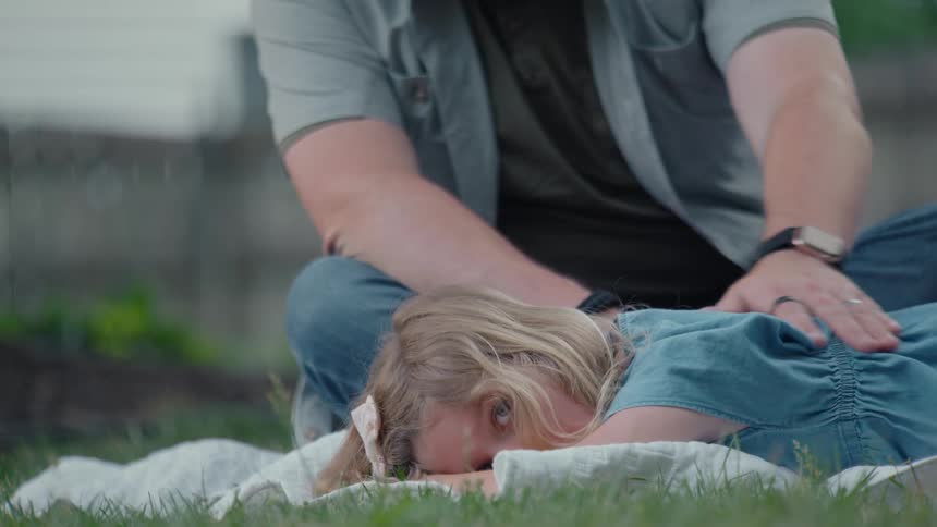 A loving father gently rubs his young daughter's back as she rests on a blanket in the grass