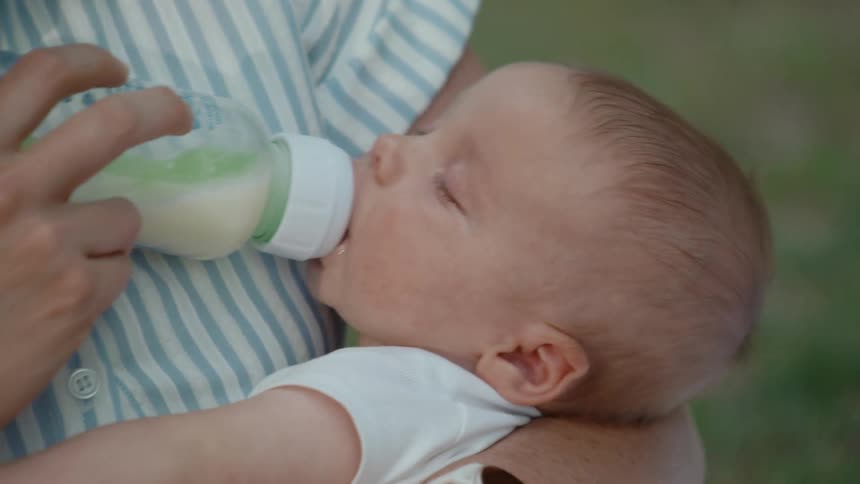 A sleepy newborn baby peacefully drinks milk from a bottle while being cradled in their mother's arms