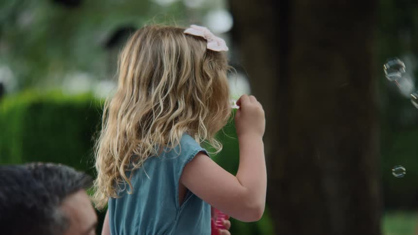 A young girl with blonde curly hair and a pink bow blows bubbles in a park in slow motion