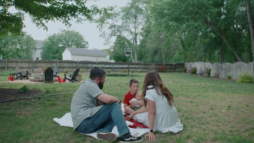 A family of five sits together on a picnic blanket in their grassy backyard on an overcast day