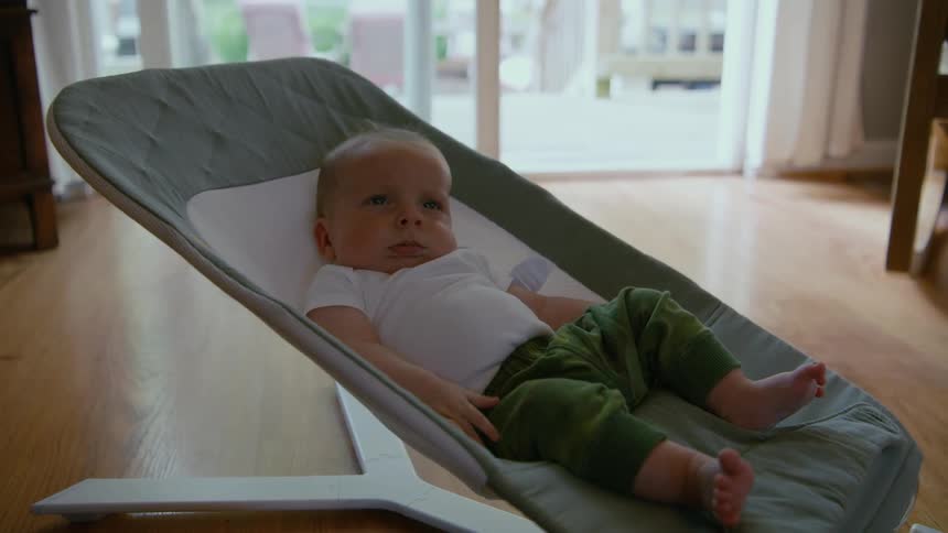 A sleepy newborn baby boy wakes up for a moment before falling back to sleep in a modern bouncer inside his home