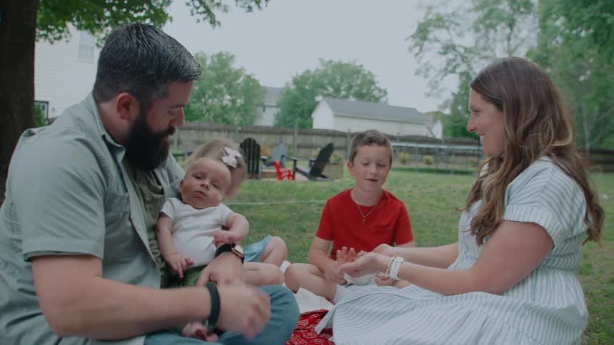 A loving family of five, including a newborn baby, shares a joyful moment playing a game together on a picnic blanket in their grassy backyard