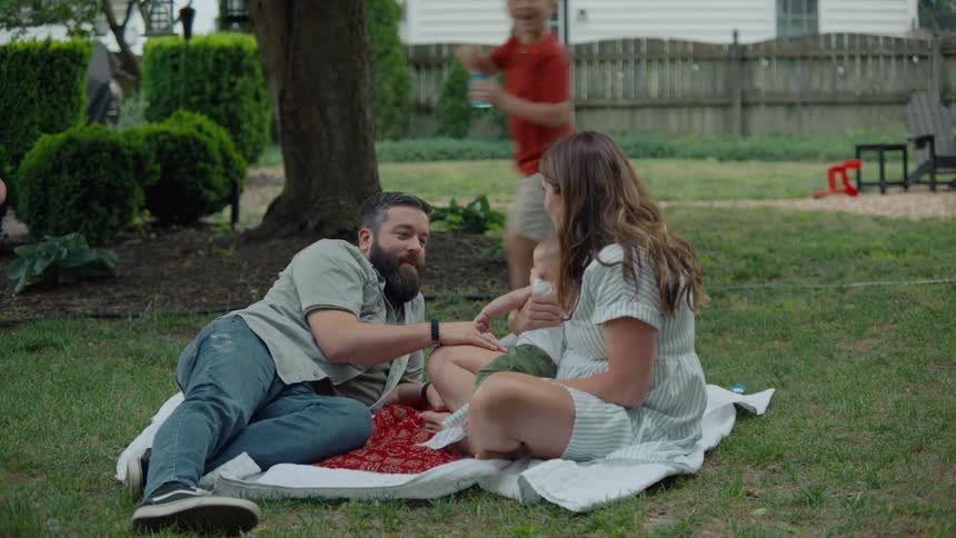 Happy parents relax on a blanket with their baby while their other children joyfully run around them playing with bubbles in the backyard